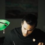 Smart Home Office - Concentrated young male wearing black turtleneck taking notes and reading books while sitting at desk with papers and bankers lamp in dark home office