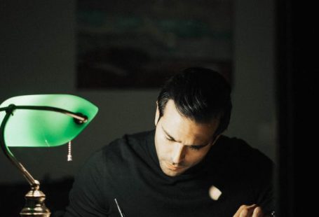 Smart Home Office - Concentrated young male wearing black turtleneck taking notes and reading books while sitting at desk with papers and bankers lamp in dark home office