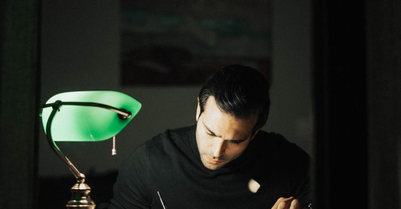 Smart Home Office - Concentrated young male wearing black turtleneck taking notes and reading books while sitting at desk with papers and bankers lamp in dark home office