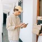 A cheerful woman receives a delivery from a person in a hallway, using a smartphone for online payment.