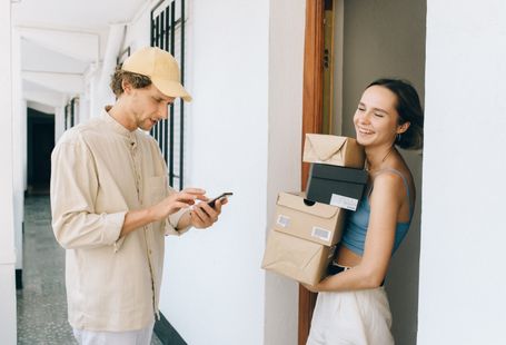 A cheerful woman receives a delivery from a person in a hallway, using a smartphone for online payment.