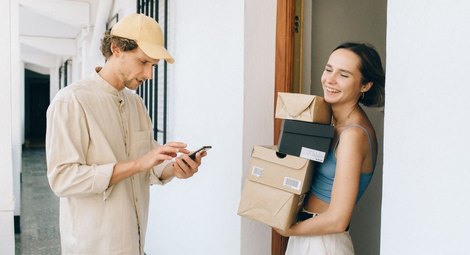 A cheerful woman receives a delivery from a person in a hallway, using a smartphone for online payment.