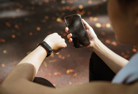 Back view of anonymous female using smartphone app and smart watch after jogging in park