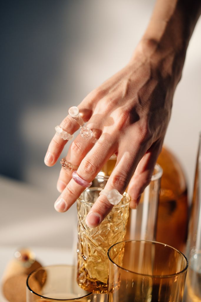 Close-up of a hand wearing multiple rings, reaching for an ornate glass under warm lighting.