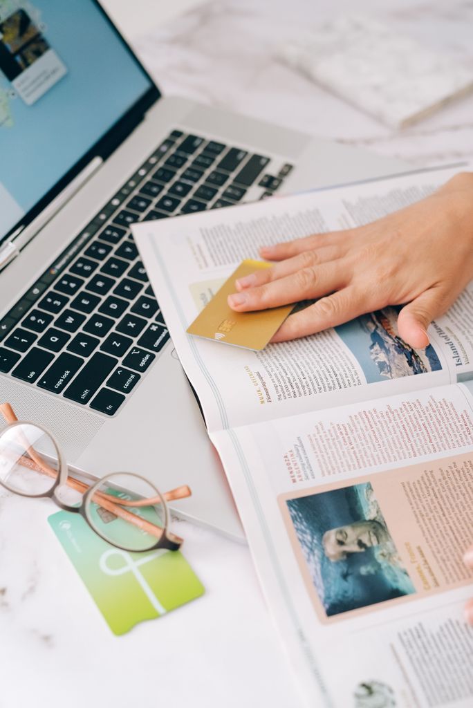 Person holding a credit card while reading a magazine next to a laptop.