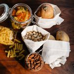 Sustainable Food Brands - Top view jars of raw pasta placed on wooden table near ECO friendly sacks with pistachios and almonds near pumpkin and potatoes