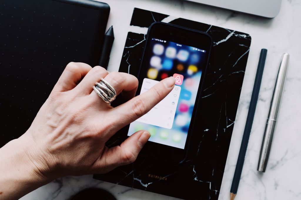 Top view of anonymous person using smartphone app tapping on screen while working with digital devices and pen and pencil placed on white marble table at home