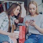 Voice Shopping -Two young women shopping together, using a smartphone in a fashion boutique with shopping bags.