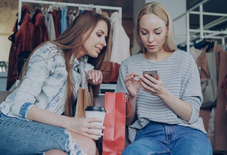 Voice Shopping -Two young women shopping together, using a smartphone in a fashion boutique with shopping bags.