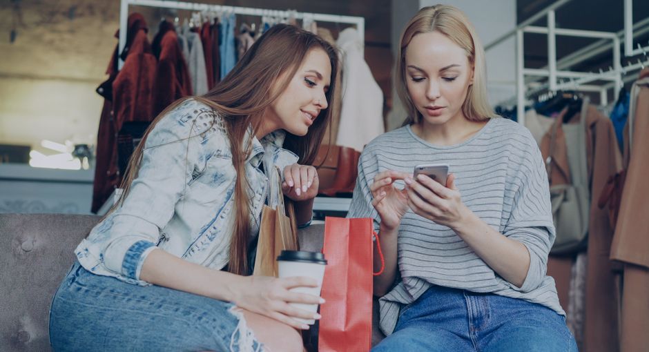 Voice Shopping -Two young women shopping together, using a smartphone in a fashion boutique with shopping bags.
